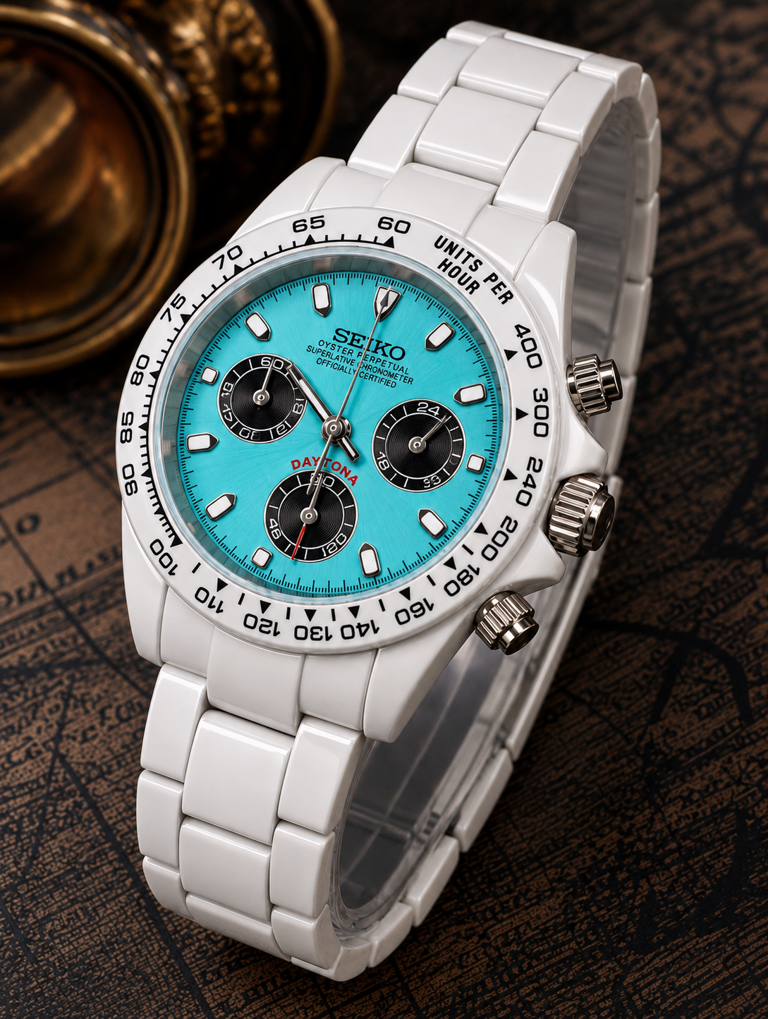 A Seiko wristwatch with a white ceramic link bracelet, a bright turquoise dial, and a black bezel, shown on a dark textured background with a brass compass prop.
